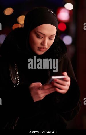 Donna d'affari musulmana Hijabi che controlla il suo telefono sulla strada urbana di notte Foto Stock