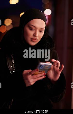 Donna d'affari musulmana Hijabi che controlla il suo telefono sulla strada urbana di notte Foto Stock