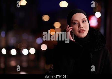 Donna d'affari musulmana Hijabi che controlla il suo telefono sulla strada urbana di notte Foto Stock