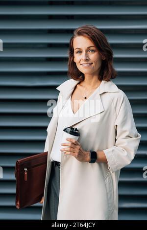 Donna d'affari di mezza età che tiene in mano una tazza di caffè e una cartella in pelle mentre si trova all'aperto Foto Stock