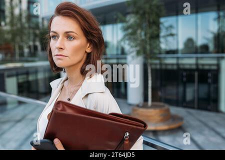 Ritratto ravvicinato di una donna d'affari di mezza età che tiene una cartella di pelle contro un edificio commerciale Foto Stock