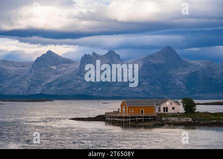 Boathouse sulla riva, la catena montuosa delle sette Sorelle dietro, l'isola di Heroy, la costa di Helgeland, Nordland, Norvegia Foto Stock