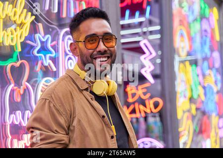 Ritratto di un giovane latino con riflessi di luce al neon sul viso. Tizio che guarda la telecamera. Hipster millenario con barba e occhiali da sole. Immagine orizzontale. Foto Stock