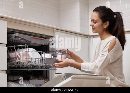 una donna attraente che scarica la tazza dalla lavastoviglie automatica aperta con utensili puliti all'interno in una moderna cucina bianca. Foto Stock