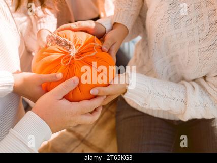 Una famiglia di quattro persone in maglioni bianchi durante le soleggiate giornate autunnali nella natura tiene in mano una grande zucca arancione fatta a mano. Concetto del giorno del Ringraziamento. Mani con Foto Stock