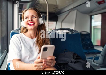 Donna felice con le cuffie che tiene il telefono cellulare tra le mani e ascolta la musica mentre viaggia per la città in autobus navetta. Foto Stock