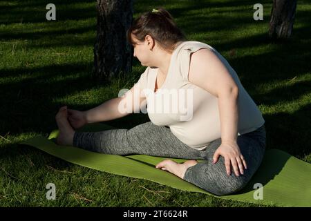 Donna di mezza età in sovrappeso che si allunga sul tappetino yoga nel parco cittadino in estate. donna grassa che fa fitness per perdere peso. Allenamento all'aperto, yoga, str Foto Stock