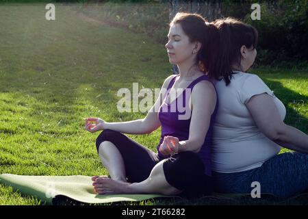 giovane donna atletica attraente e donna in sovrappeso che meditano nella natura, seduti a schiena. Salute mentale, stile di vita sano, yoga all'aperto. Zen, Foto Stock