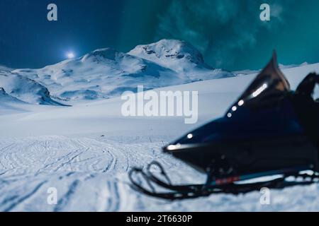 Foto panoramica molto ravvicinata del lato anteriore della motoslitta intenzionalmente sfocata con montagne notturne e luci verdi settentrionali nel cielo stellato. Backg Foto Stock