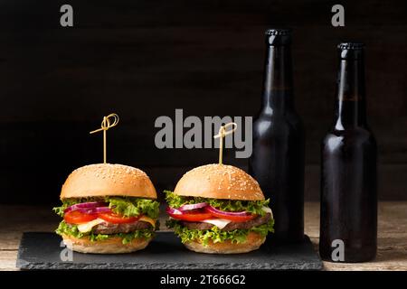 Due hamburger, patatine fritte, birra in bottiglie scure su fondo di legno. Fast food, hamburger fatti in casa Foto Stock