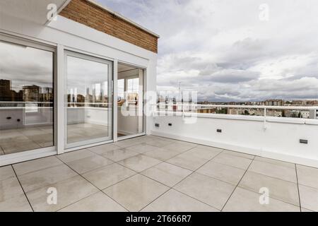 Terrazza con pavimento in gres bianco, grande finestra con tre porte in alluminio e vetro bianco, pareti combinate in mattoni e intonaco bianco, vista della città Foto Stock