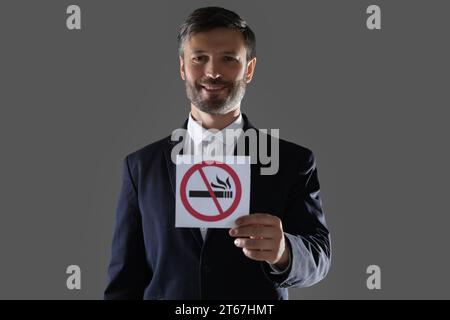 Uomo che tiene una carta con un cartello di divieto di fumo su sfondo grigio Foto Stock