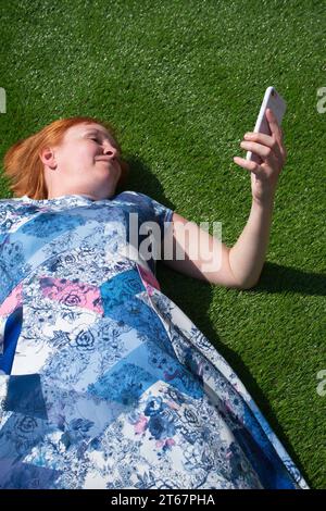 Una donna dai capelli rossi che indossa un abito blu a motivi geometrici, sdraiata a faccia in su e sorridente sull'erba artificiale al sole, guardando il suo telefono Foto Stock