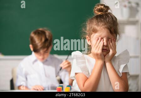 Una ragazza intelligente della scuola grida. Studentessa in uniforme coprendo il suo viso con il braccio piangendo triste di bullismo a scuola in piedi di fronte alla lavagna Foto Stock