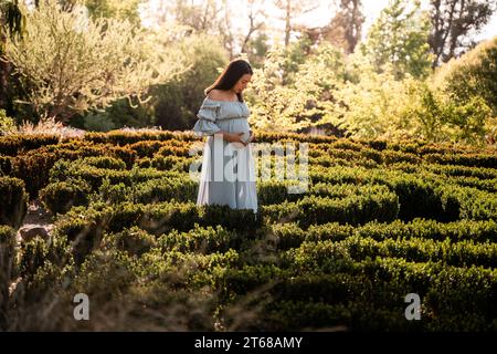 Una donna latina incinta in un vestito in piedi in un lussureggiante parco illuminato dal sole, le mani cullano dolcemente la pancia Foto Stock