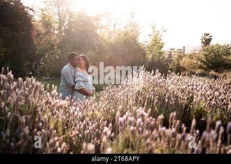 Una donna latina incinta e una coppia di uomini africani in piedi in un parco lussureggiante e luminoso illuminato dal sole Foto Stock