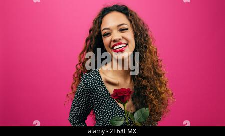 La bella donna multietnica puzza di rosa rossa, ridendo e sorridendo, studio rosa Foto Stock