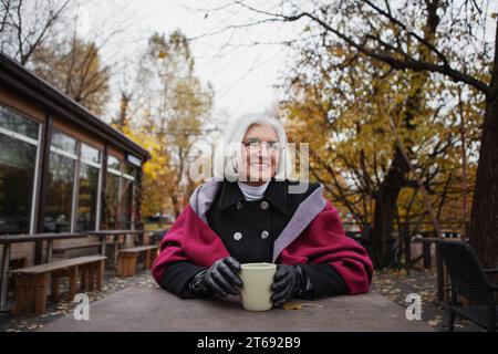 Donna anziana sorridente con i capelli grigi seduta al tavolo del caffè sulla strada nel parco autunnale con una tazza di tè o caffè, indossando un cappotto e indossando una comoda confezione Foto Stock