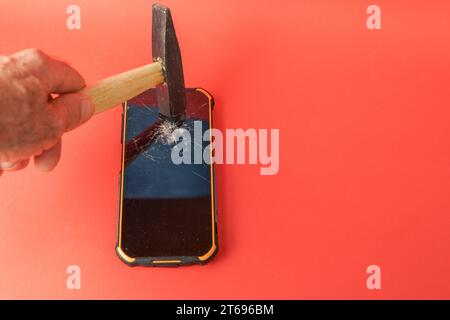 la mano di un uomo con un martello che rompe lo schermo di un telefono cellulare su uno sfondo rosso Foto Stock