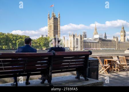 Due uomini siedono a parlare su un posto a sedere con il Palazzo di Westminster, luogo di incontro del Parlamento del Regno Unito, sullo sfondo Foto Stock