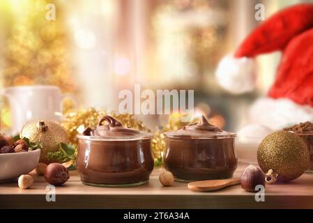 Due vasetti di vetro pieni di crema di cacao con nocciole con nocciole intorno a un tavolo di legno nella sala da pranzo della casa con decorazioni natalizie. Anteriore V Foto Stock