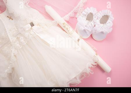 Abito da battesimo per bambini su sfondo rosa con croce, candela e scarpe per bambini Foto Stock
