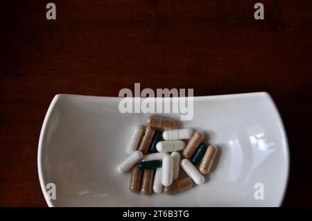 Una piccola piastra bianca contiene molte pillole e capsule di diversi colori. Una ciotola bianca di varie pillole colorate e capsule isolate su dorso marrone Foto Stock