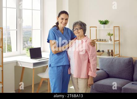 Giovane infermiera sorridente che assiste e abbraccia la paziente anziana a casa. Foto Stock