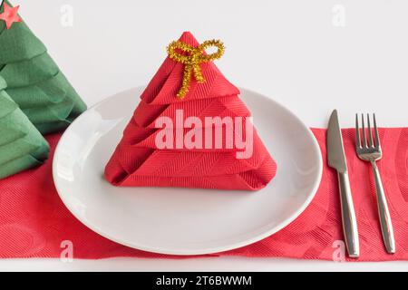 Tovagliolo di carta di colore rosso piegato a forma di albero di Natale su piatto bianco con posate Foto Stock