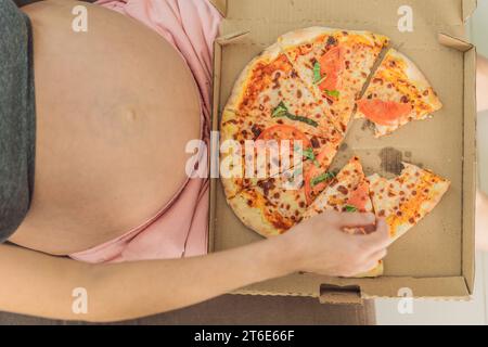 Una donna incinta si diverte a gustare una fetta di pizza, assaporando un momento di indulgenza e soddisfacendo il suo desiderio di una delizia deliziosa e confortante. Entusiasta Foto Stock