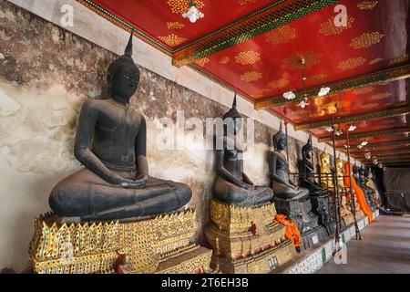 Una fila infinita di statue di Buddha in oro e nero fiancheggiano un portico che circonda il tempio, alcune figure sono in fase di ristrutturazione. A Wat Suthat Thepwa Foto Stock