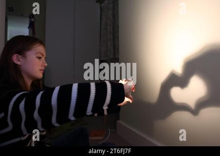 Ragazza adolescente che crea un cuore ombra con le mani e le dita con una lampada sulla parete Foto Stock