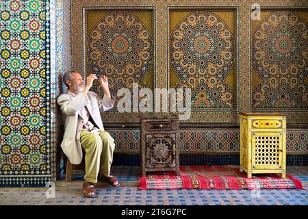 Un uomo dall'aspetto distinto pulisce gli occhiali mentre si siede davanti A Una parete a mosaico dai motivi spettacolari Foto Stock