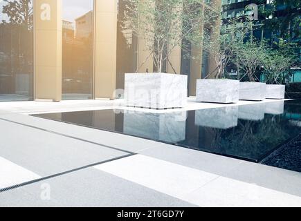 Bellissimo giardino di fronte a un edificio commerciale, una fila di grandi vasi per piante, un piccolo laghetto e un bel pavimento, luce calda. Il concetto di edificio aziendale. Foto Stock