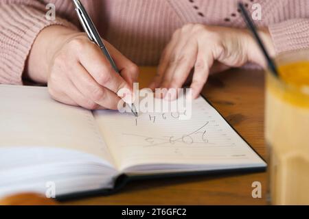 La mano di una donna 2024. Routine di Capodanno, risoluzioni alla scrivania. Cafe, interni del ristorante sullo sfondo. elenco obiettivi 2024 con notebook, tazza di caffè su w Foto Stock