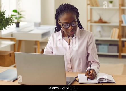 Donna d'affari afroamericana seria che lavora su un computer portatile e prende appunti in ufficio. Foto Stock