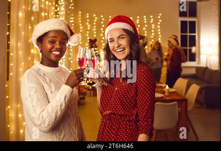 Due donne interrazziali con cappello di Babbo Natale e bicchieri di vino rosso dicono brindisi per felice anno nuovo. Foto Stock