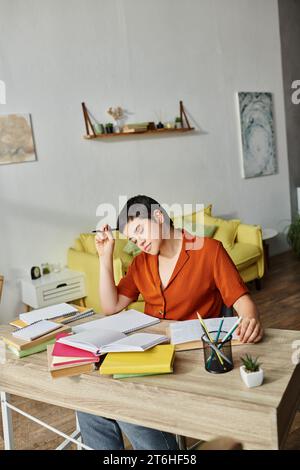 immagine verticale di uno studente stanco che lavora sodo seduto alla scrivania con una penna in mano per leggere un libro di testo Foto Stock