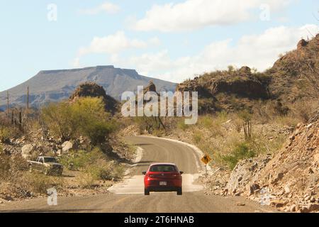 Una vivace auto rossa che viaggia lungo una tortuosa strada sterrata circondata da maestose montagne e dal deserto sbiancato dal sole Foto Stock