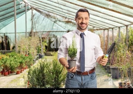 Un uomo d'affari sorridente che tiene un piccolo albero in un vaso e una vanga in un giardino Foto Stock