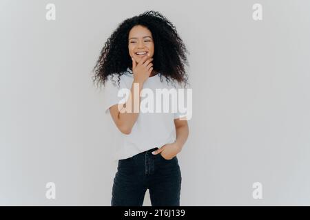 Una giovane donna che ride con una t-shirt bianca casual e un paio di jeans, mano sul mento, con un'espressione gioiosa Foto Stock