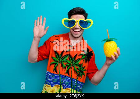 Foto di un uomo di buon umore con una elegante camicia stampata vestita di capelli in divertenti occhiali tenere le ananas bere dire ciao isolato su sfondo di colore blu Foto Stock