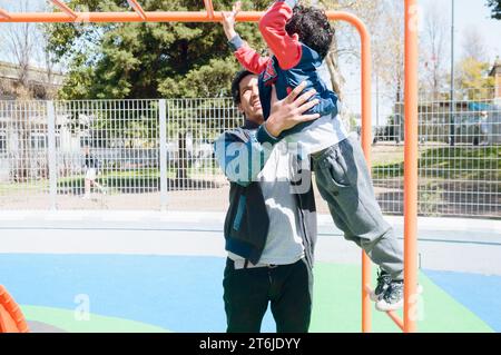 giovane padre latino venezuelano con afro, che aiuta suo figlio a giocare nei giochi dei parchi pubblici a mezzogiorno, insieme nel fine settimana a buenos aires Foto Stock