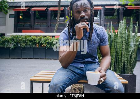 Vista frontale del giovane afro con dreadlock e barba di etnia africana, indossando abiti informali seduti, guardando la macchina fotografica tenendo seriamente la tazza Foto Stock