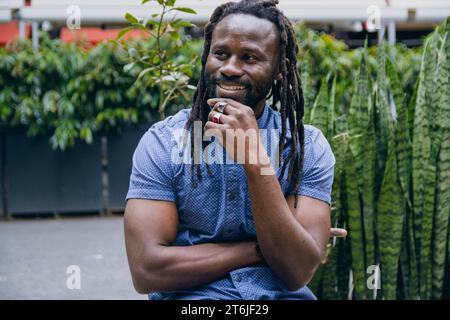 Ritratto di bel giovane africano con dreadlock e barba, indossa abiti casual blu, siediti all'aperto a ridere e a guardare a parte con attenzione Foto Stock