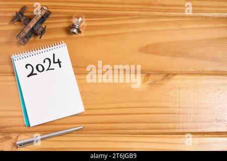 Vista dall'alto dell'elenco vuoto 2024 sul notebook su un tavolo di legno con aereo in miniatura, piccola lampadina e penna. Obiettivi e risoluzioni per il nuovo anno, nuovo inizio a. Foto Stock