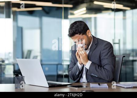 Un uomo starnutisce in un tessuto in un luogo di lavoro all'interno di un ufficio, un uomo d'affari è malato con il naso che cola, lavora in un luogo di lavoro con allergie, usa un notebook al lavoro. Foto Stock