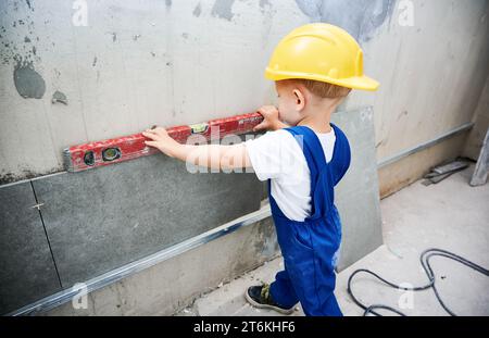 Lavoratore edile per bambini che controlla la superficie della parete con una livella a bolla d'aria nell'appartamento. Capretto in tute da lavoro che utilizza uno strumento di livellamento durante la preparazione della parete per lavori di riparazione a casa durante la ristrutturazione. Foto Stock