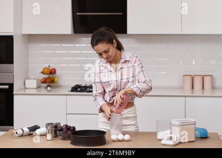 Donna di casa felice che prepara la torta, aggiungendo gli ingredienti alla ciotola nella cucina moderna Foto Stock