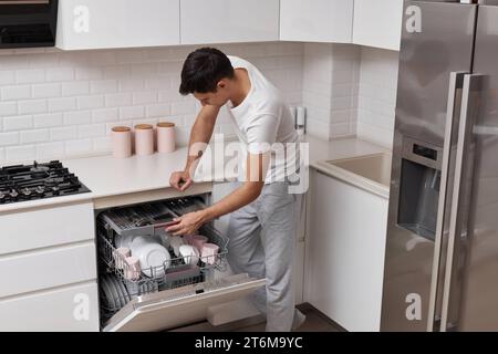 bell'uomo positivo che mette piatti in lavastoviglie in cucina Foto Stock